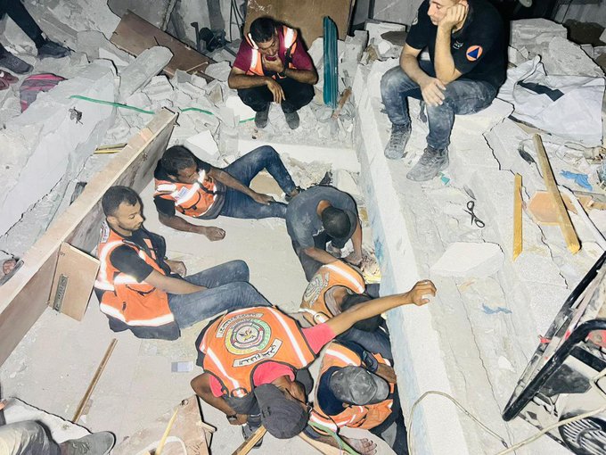Rescue workers in orange vests with reflective stripes sit and work amidst rubble and debris. The scene shows a destroyed building with scattered concrete, wood, and household items. One worker in a black shirt sits on the edge, observing the area.