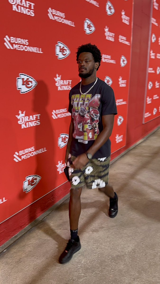 Tyquan Thornton walking in a hallway. He is wearing a black T-shirt with a graphic of a football player, camouflage shorts with white floral patterns, and black sneakers. The background features red walls with Kansas City Chiefs logos and DraftKings branding.