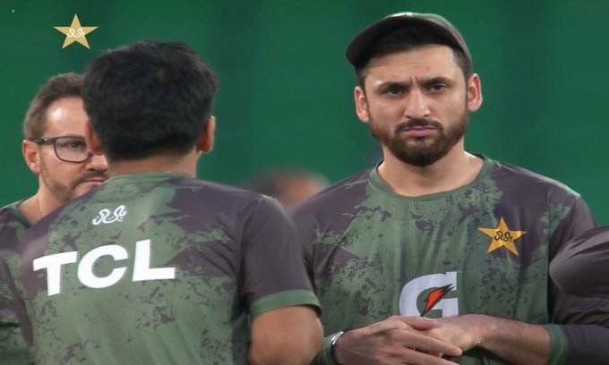 Salman Ali Agha wearing a green cricket jersey with a star emblem and "TCL" text on the back. Another person in a similar jersey faces away, showing the same design. Both individuals stand on a green field, with a Gatorade logo visible on Salman Ali Agha\'s jersey.