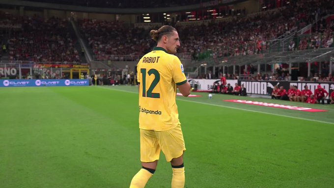 Adrien Rabiot wearing a yellow AC Milan jersey with the number 12 and his name on the back, standing on a soccer field. A large crowd is visible in the stadium stands. The field is green with visible advertisements along the sidelines.