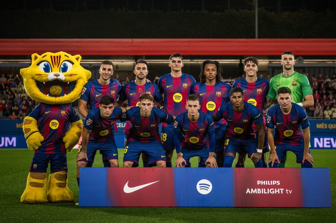 A group of FC Barcelona soccer players in blue and red striped jerseys standing and kneeling on a field. A mascot in a yellow and red tiger costume stands to the left. A banner with Nike, Spotify, and Philips Ambilight TV logos is visible in front.