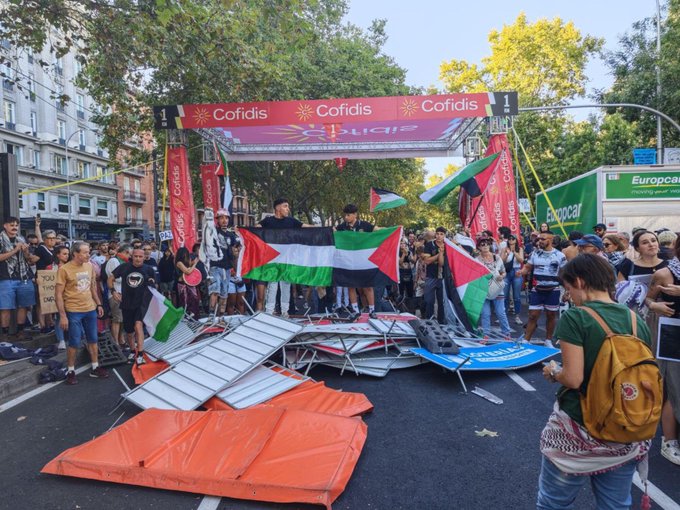 A street scene with a collapsed finish line structure, including metal barriers and orange fabric, lying on the road. People hold Palestinian flags and gather around the structure, some standing on it. Banners with "Cofidis" and "La Vuelta" are visible above the scene, along with trees and buildings in the background.