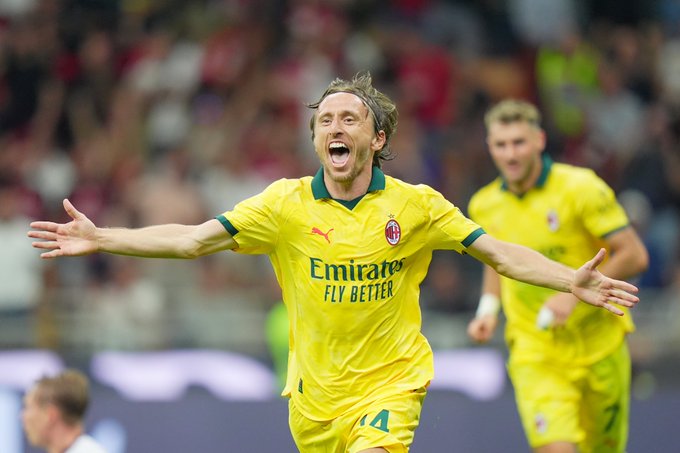 Luka Modrić wearing a yellow AC Milan jersey with the number 14 and the "Emirates Fly Better" logo. He has his arms outstretched, celebrating on a soccer field. Another player in a similar yellow AC Milan jersey is visible in the background.
