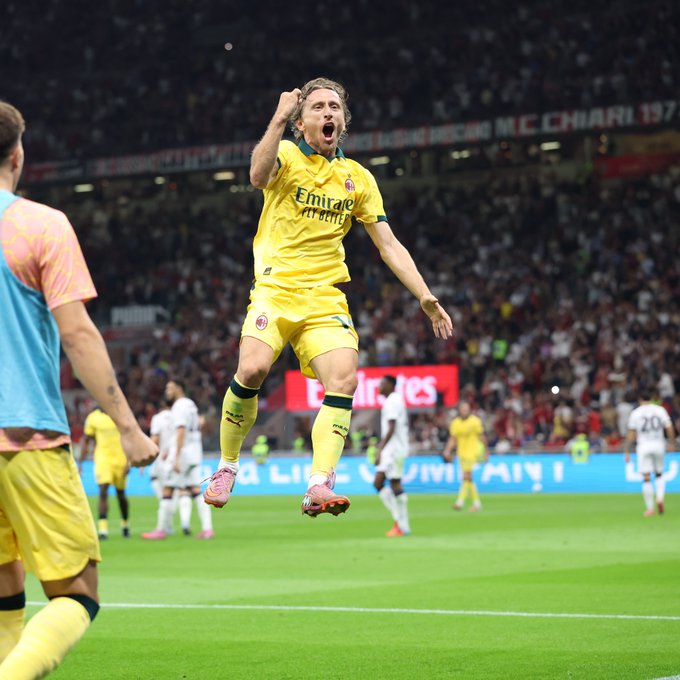 A soccer player in a yellow and black AC Milan jersey celebrating on a field, arms raised, mid-jump. The stadium is filled with spectators, and other players in white and yellow uniforms are visible in the background. The player wears pink cleats and black socks with yellow accents.