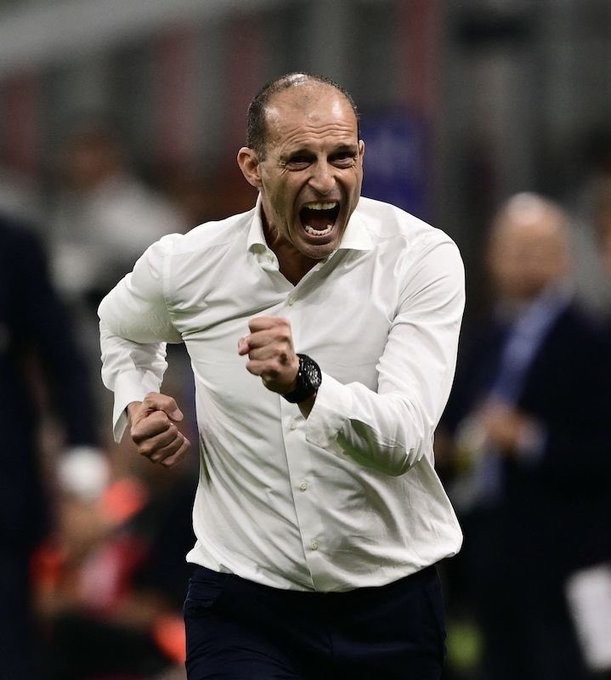 Max Allegri wearing a white dress shirt and dark pants, running with a clenched fist and an open mouth, appearing to celebrate. A blurred crowd and stadium seating are visible in the background.