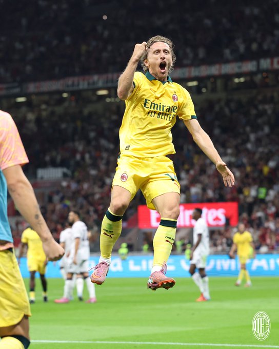 A soccer player in a yellow AC Milan jersey with "Emirates" text, celebrating with one arm raised and mouth open. The player wears yellow socks and pink cleats, jumping on a green field. Other players in similar yellow jerseys and white jerseys are visible in the background, along with a stadium crowd.