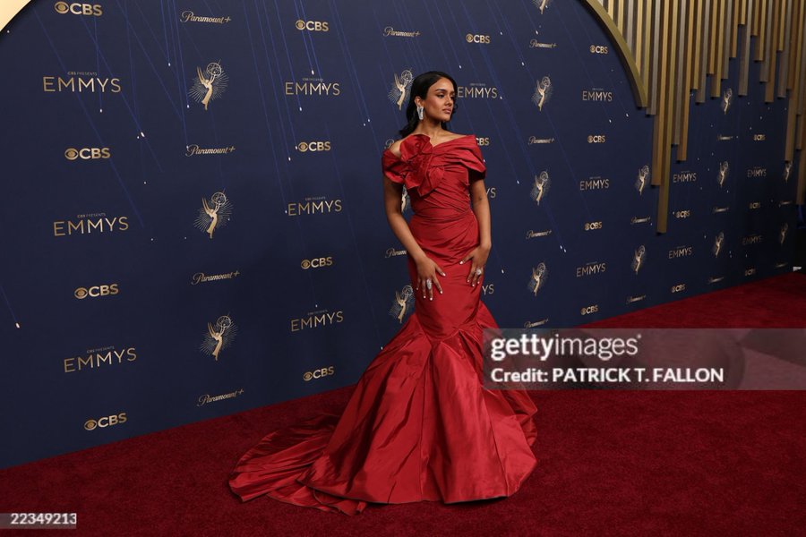 A woman in a red, floor-length gown with off-shoulder design and ruffled details, standing on a red carpet. She has long, dark hair and wears dangling diamond earrings. The backdrop features repeated "Emmys" and "CBS" logos.