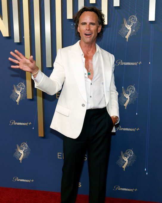 Walton Goggins stands on a red carpet, wearing a white blazer, white shirt, and black pants. He has a necklace and bracelets, with one hand raised and the other touching his chin. The backdrop features Paramount and Emmy logos with gold and blue designs.