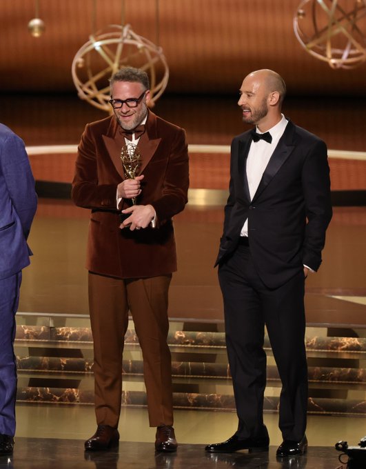 Three men on a stage at an awards event. One man in a brown velvet suit holds an Emmy statuette, another in a black tuxedo with a bow tie stands beside him, and a third in a blue suit is partially visible. The background features golden and blue decor with spherical ornaments. The setting suggests a formal ceremony.