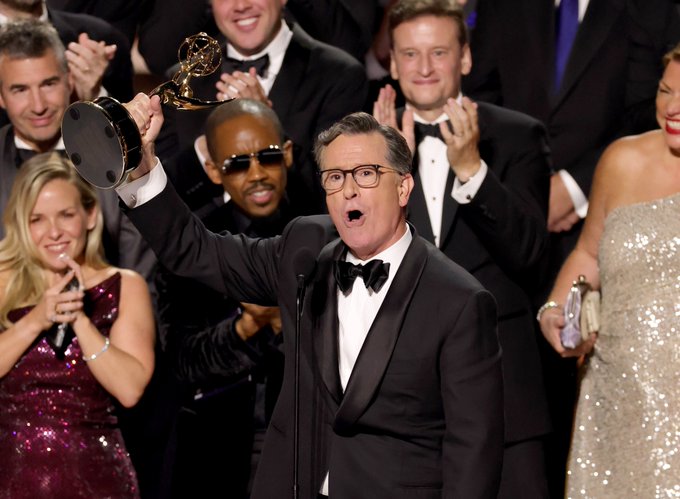 Stephen Colbert holding an Emmy Award trophy above his head, wearing a black tuxedo with a bow tie. He stands on a stage surrounded by people in formal attire, some clapping. The background shows a crowd in suits and dresses.