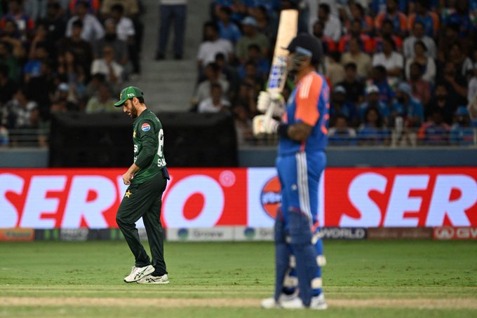 Salman Ali Agha in a green cricket uniform walking on a field, holding a bat. An opposing player in an orange and blue uniform stands nearby, also holding a bat. A crowd is visible in the background, and a digital advertisement board with the word "SER" is present.