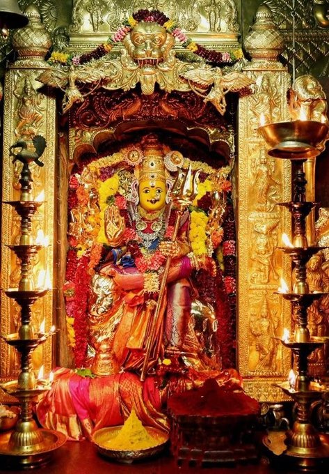 A golden statue of Kanakadurga Devi adorned with vibrant red and yellow garments and garlands. The statue holds a trident and is seated within an ornate, intricately decorated temple shrine. Oil lamps and offerings are placed around the statue, with a backdrop of detailed carvings and decorations.