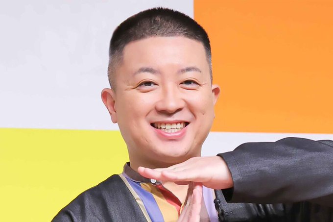 A person with short dark hair, wearing a black jacket and colorful shirt, smiling and raising a hand in a gesture. The background features a colorful design with yellow and orange sections.