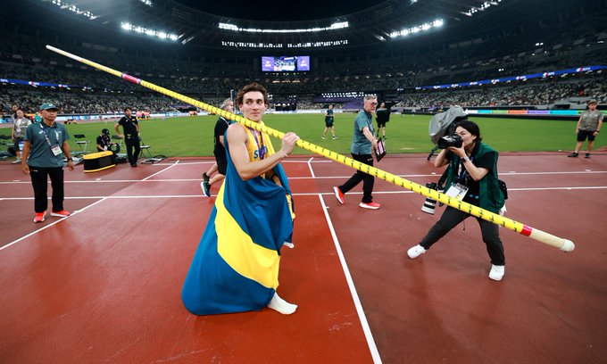 Armand Duplantis sosteniendo un palo de salto con pértiga en la pista de un estadio de atletismo. Viste un uniforme azul y amarillo. Una persona con una cámara está cerca, capturando el momento. El estadio está lleno de espectadores y luces brillantes.