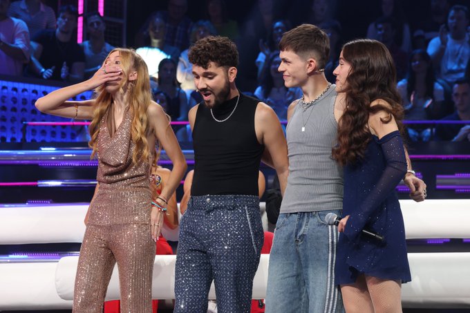 Guille Milkyway, Lucia Casani, and Cristina stand on a brightly lit stage with a live audience in the background. Guille Milkyway wears a sleeveless black top and sparkly pants. Lucia Casani is dressed in a shimmering bronze outfit, covering her mouth with her hand. Cristina wears a blue dress with long sleeves. Another person in a gray top and jeans stands among them.