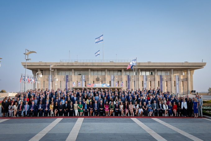 A large group of 250 US state legislators posing for a group photo in front of a building. The building has multiple Israeli flags and other national flags visible on poles. The legislators are dressed in formal attire, including suits and dresses, and are arranged in several rows on a paved area with a red carpet.