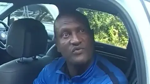 Hadush Kebatu sitting inside a car, wearing a blue shirt with black stripes. The car has black seats and a visible steering wheel, with greenery seen through the window.