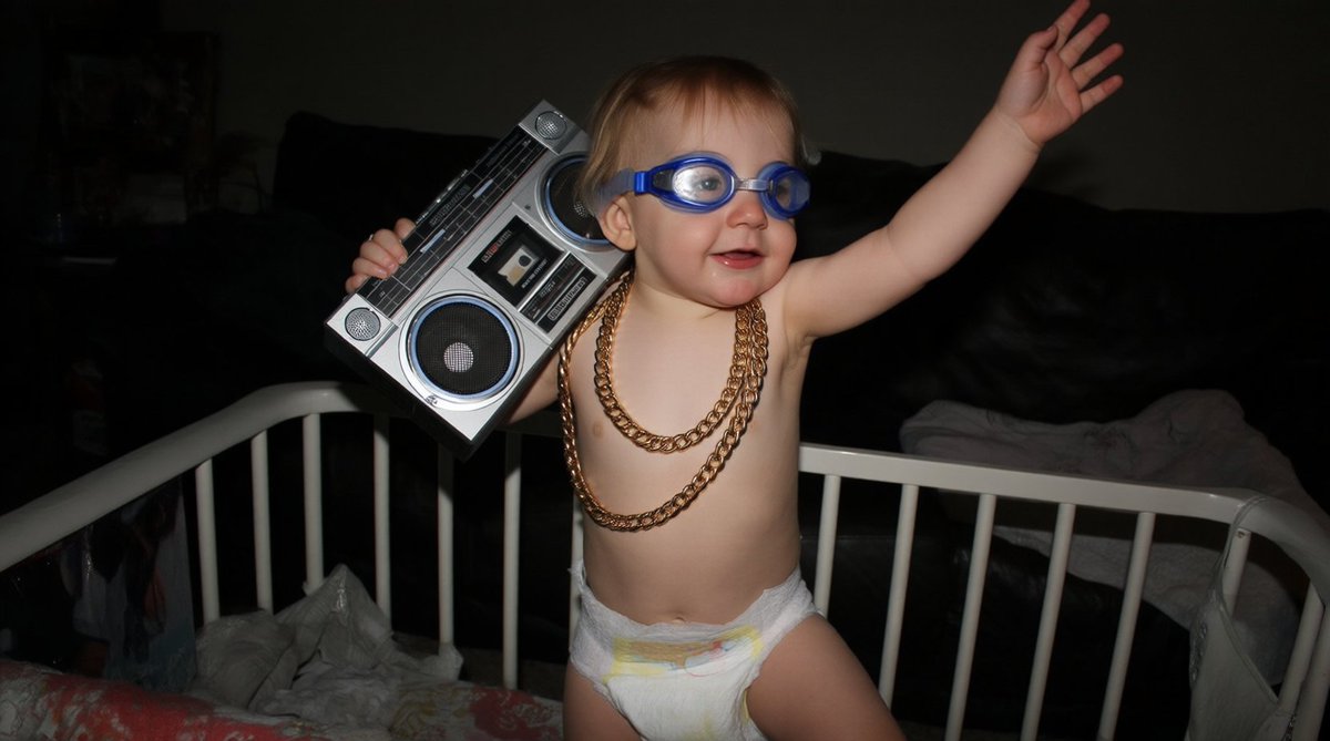 Flikr photo from 2012 featuring a baby boy wearing a diaper and swim goggles, the baby has thick gold chains around its neck and a large boombox on its shoulder, the baby is smiling and looks happy, it is standing in its crib, seemingly dancing in a dynamic pose