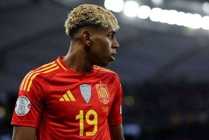 Lamine Yamal wearing a red Spain national team soccer jersey with yellow accents. The jersey has the number 19 on it and features the Spain crest. His hair is blonde and styled in short curls.