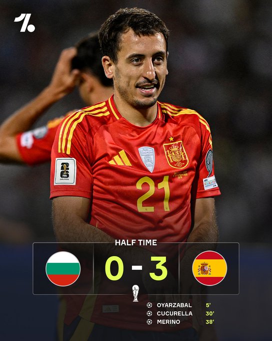 A man wearing a red Spain national team soccer jersey with the number 21 on it, standing on a field. The jersey has yellow stripes on the shoulders and the Spain crest on the chest. Another player in a similar jersey is partially visible in the background. A scoreboard overlay shows Bulgaria 0-3 Spain at half time, with goals by Oyarzabal at 5 minutes, Cucurella at 30 minutes, and Merino at 38 minutes.