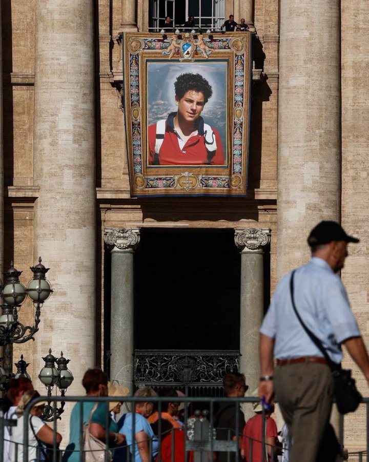 A large framed portrait of Carlo Acutis displayed on the facade of a historic stone building with columns and an arched entrance. The portrait shows Carlo Acutis wearing a red shirt and white collar. People are visible in the foreground, some standing near ornate street lamps.
