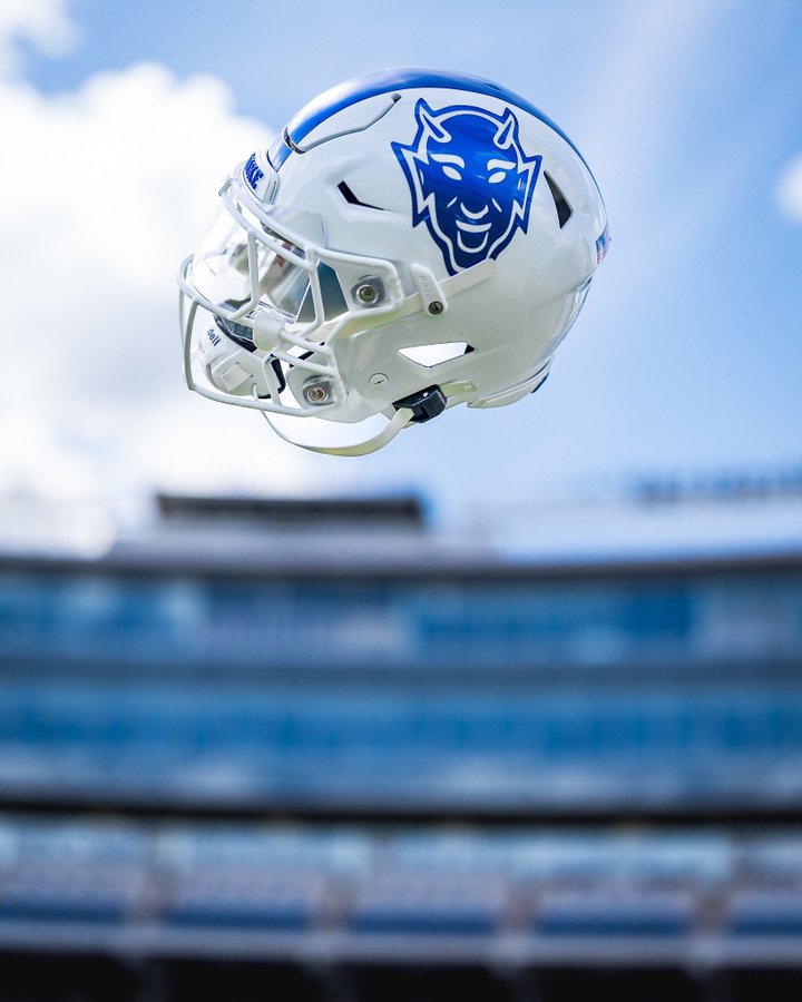 A white football helmet with a blue devil logo on the side, mid-air against a backdrop of a stadium with blue seating and a partly cloudy sky.