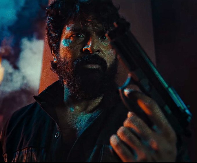 Sivakarthikeyan holding a gun in a dark setting. He has a beard and is wearing a black jacket. Smoke and colorful lighting are visible in the background.