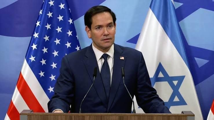 Marco Rubio stands at a podium, wearing a dark suit, white shirt, and tie. Behind him are the American flag and the Israeli flag. The setting appears formal, with a blue background.