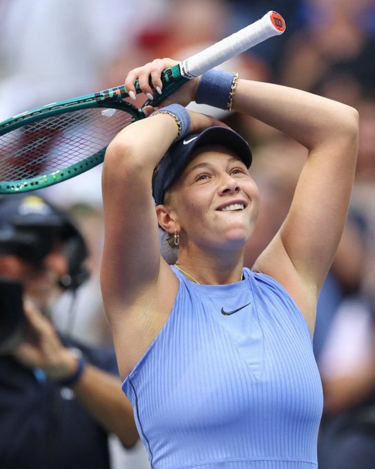 Amanda Anisimova holding a tennis racket above her head with both hands. She is wearing a blue sleeveless top, a navy cap, and wristbands. Her expression shows a smile, and she appears to be on a tennis court with a crowd in the background.