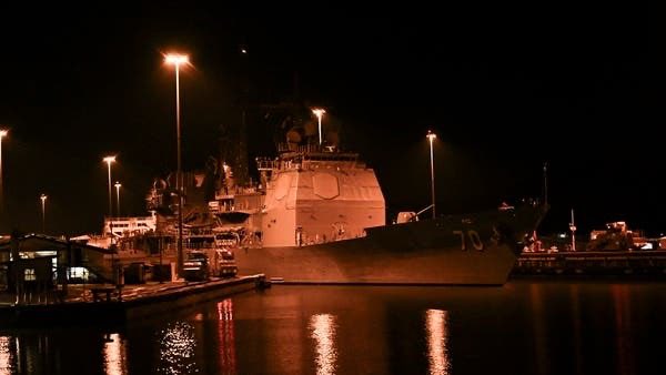Un gran barco de la Armada atracado en un puerto por la noche. El barco está iluminado por farolas, proyectando reflejos en el agua. El buque tiene una estructura prominente con equipo de radar y un número de casco visible.