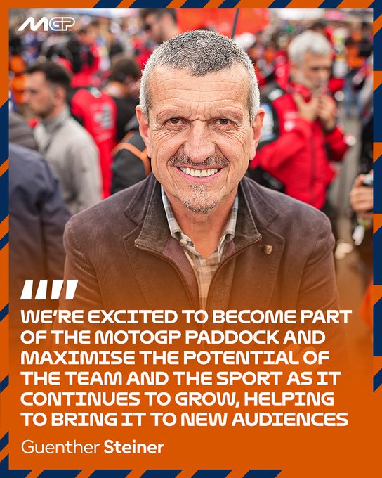 Guenther Steiner stands in a crowd, wearing a brown jacket over a checkered shirt. Red and orange banners are visible in the background, along with people in red jackets. Text overlay reads, "WE\'RE EXCITED TO BECOME PART OF THE MOTOGP PADDOCK AND MAXIMISE THE TEAM AND THE SPORT AS IT CONTINUES TO GROW, HELPING TO BRING IT TO NEW AUDIENCES Guenther Steiner."