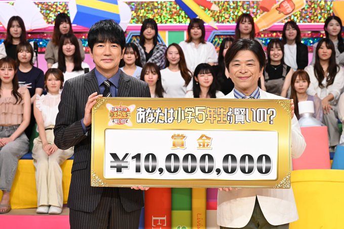 Shinsuke Hatabuki and another person holding a large golden sign displaying "¥10,000,000" and Japanese text. They are standing on a colorful set with bright yellow and blue elements, surrounded by a group of people seated in the background. The setting includes a festive atmosphere with confetti-like decorations.