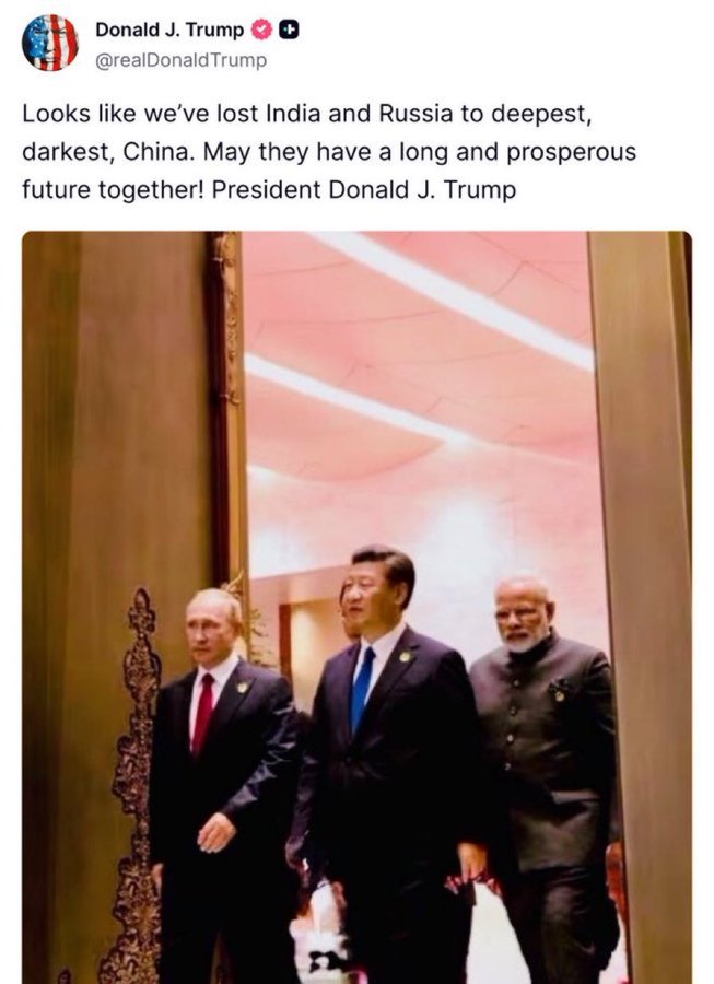 A photo of Vladimir Putin, Xi Jinping, and Narendra Modi walking together in a grand hallway with ornate decor. Donald J. Trump is not visible in the image. The hallway has a pinkish hue and features a decorative archway and columns.