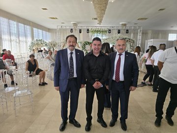 A group of men in suits and formal attire standing together in a decorated indoor venue. Some wear red ties, others blue or black, with one in a black shirt. A chandelier hangs above, and floral arrangements decorate the background. Banners display "Kıbrıs Türk Memur-Sen" and Turkish flags, with text reading "8. Olağan Genel Kuruluna" and portraits of individuals.