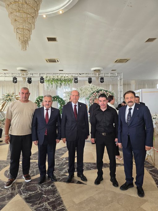 A group of men in suits and formal attire standing together in a decorated indoor venue. Some wear red ties, others blue or black, with one in a black shirt. A chandelier hangs above, and floral arrangements decorate the background. Banners display "Kıbrıs Türk Memur-Sen" and Turkish flags, with text reading "8. Olağan Genel Kuruluna" and portraits of individuals.