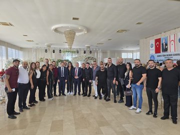 A group of men in suits and formal attire standing together in a decorated indoor venue. Some wear red ties, others blue or black, with one in a black shirt. A chandelier hangs above, and floral arrangements decorate the background. Banners display "Kıbrıs Türk Memur-Sen" and Turkish flags, with text reading "8. Olağan Genel Kuruluna" and portraits of individuals.