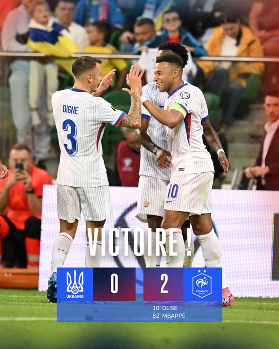Three soccer players in white uniforms with blue and red accents celebrating on a field. One player, wearing the number 3 jersey with "Digne" on the back, high-fives another player in a number 10 jersey. A scoreboard shows Ukraine 0, France 2, with Kylian Mbappé listed as a goal scorer. Spectators are visible in the background.