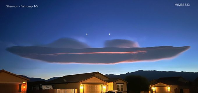 A massive, flat lenticular cloud stretches across the twilight sky, glowing at the edges with a reddish hue. The cloud hovers above mountains, resembling a giant craft. Below, houses with lit outdoor lights are visible in Pahrump, Nevada, against a backdrop of distant hills.