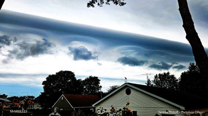 A dramatic sky with a large, horizontal cloud formation resembling a wall over Delphos, Ohio. The clouds have unusual, non-random patterns, including wave-like shapes. Below the sky, houses with sloped roofs, trees, and a yard with sunflowers are visible. A watermark reads "Tara Thrift - Delphos, Ohio."