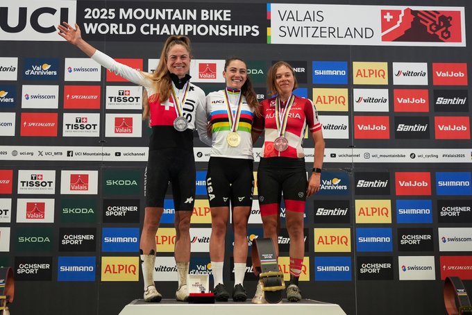 Kate Courtney, Anna Weinbeer, and Mona Mitterwallner stand on a podium. Kate Courtney wears a gold medal and a USA jersey, Anna Weinbeer wears a silver medal and a Swiss jersey, and Mona Mitterwallner wears a bronze medal and an Austrian jersey. They are dressed in cycling gear, standing on a platform with banners displaying UCI, Valais, and sponsor logos like Tissot and Alpiq.