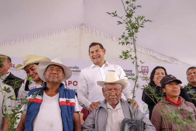 Several people wearing cowboy hats and casual clothing, including a man in a white shirt and hat, stand together holding young trees with visible roots. Some individuals have dirt on their hands and clothes, suggesting planting activity. A banner in the background displays the word "Puebla" and a logo.