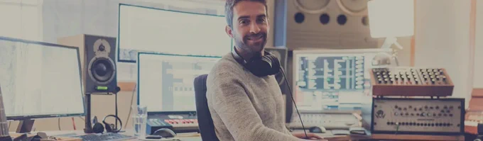 Person sitting at a desk in a professional music studio, surrounded by audio equipment, smiling at the camera.
