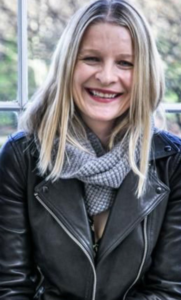A woman with long blonde hair wearing a black leather jacket and a gray patterned scarf, smiling in front of a window with greenery visible outside.