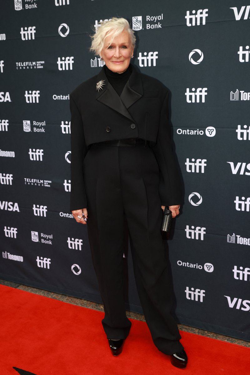 Glenn Close standing on a red carpet. She wears a black outfit with a cropped jacket, matching pants, and black shoes. A black clutch is in her hand, and a brooch is pinned to her jacket. The backdrop displays multiple TIFF logos, Royal Bank, Ontario, and Visa logos.