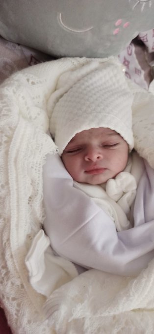 A sleeping baby wrapped in white blankets and wearing a white hat. Stacked beside the baby are packages of diapers and containers of baby formula, including brands like BabyDream, Sahha, and Nolfix. A person in a black headscarf and patterned clothing is holding the items.