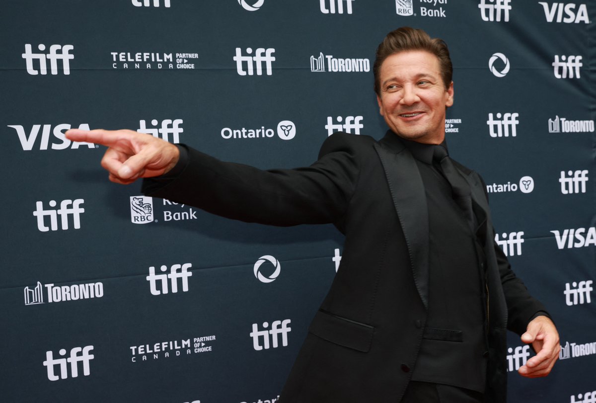Jeremy Renner stands on a red carpet, pointing with his right hand. He wears a black suit and black shirt. The backdrop displays logos for TIFF, Telefilm Canada, Ontario, Visa, and Toronto, with repeated "TIFF" text.