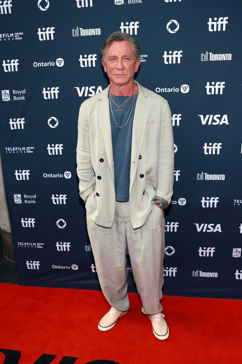 Daniel Craig stands on a red carpet, wearing a light beige suit, blue shirt, necklace, and white sneakers. He has hands in pockets, positioned in front of a backdrop with TIFF, Ontario, Royal Bank, and Visa logos.