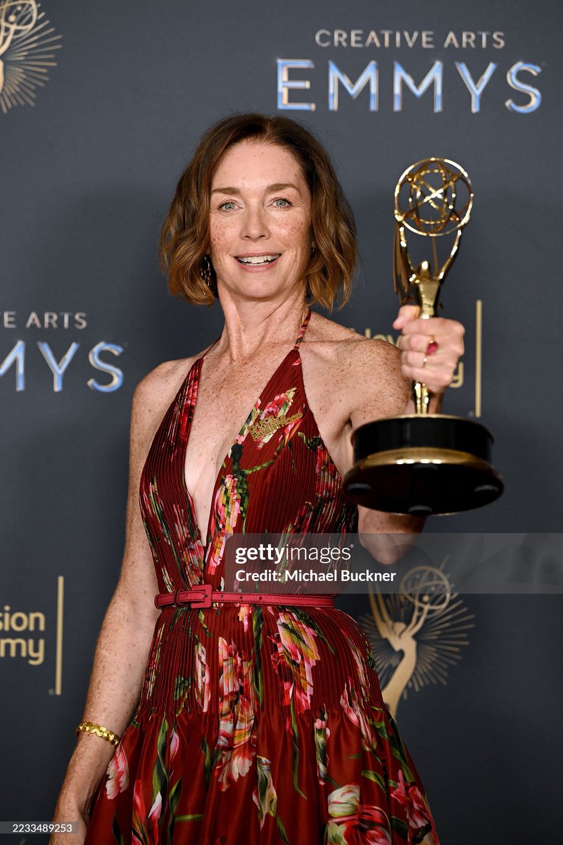 Julianne Nicholson holding an Emmy statuette. She is wearing a red floral dress with a deep V-neck and a red belt. The background features a dark backdrop with the "Creative Arts Emmys" text and Emmy logos. A Getty Images watermark is visible.
