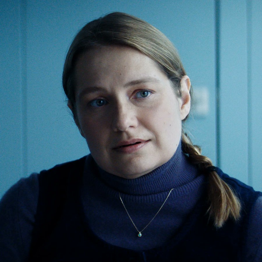 Merritt Wever with light brown hair in a ponytail, wearing a dark turtleneck and a necklace with a small pendant, standing against a blue background.