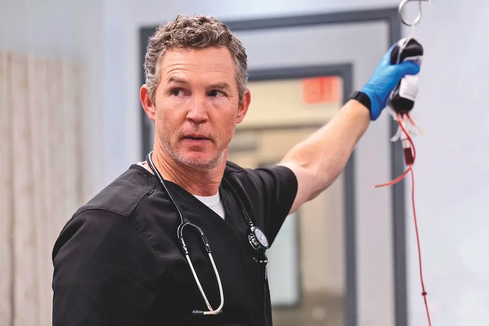 Shawn Hatosy wearing a black scrub top and white undershirt, holding a blood bag with red liquid in a hospital setting. He has a stethoscope around his neck and blue gloves on his hands.